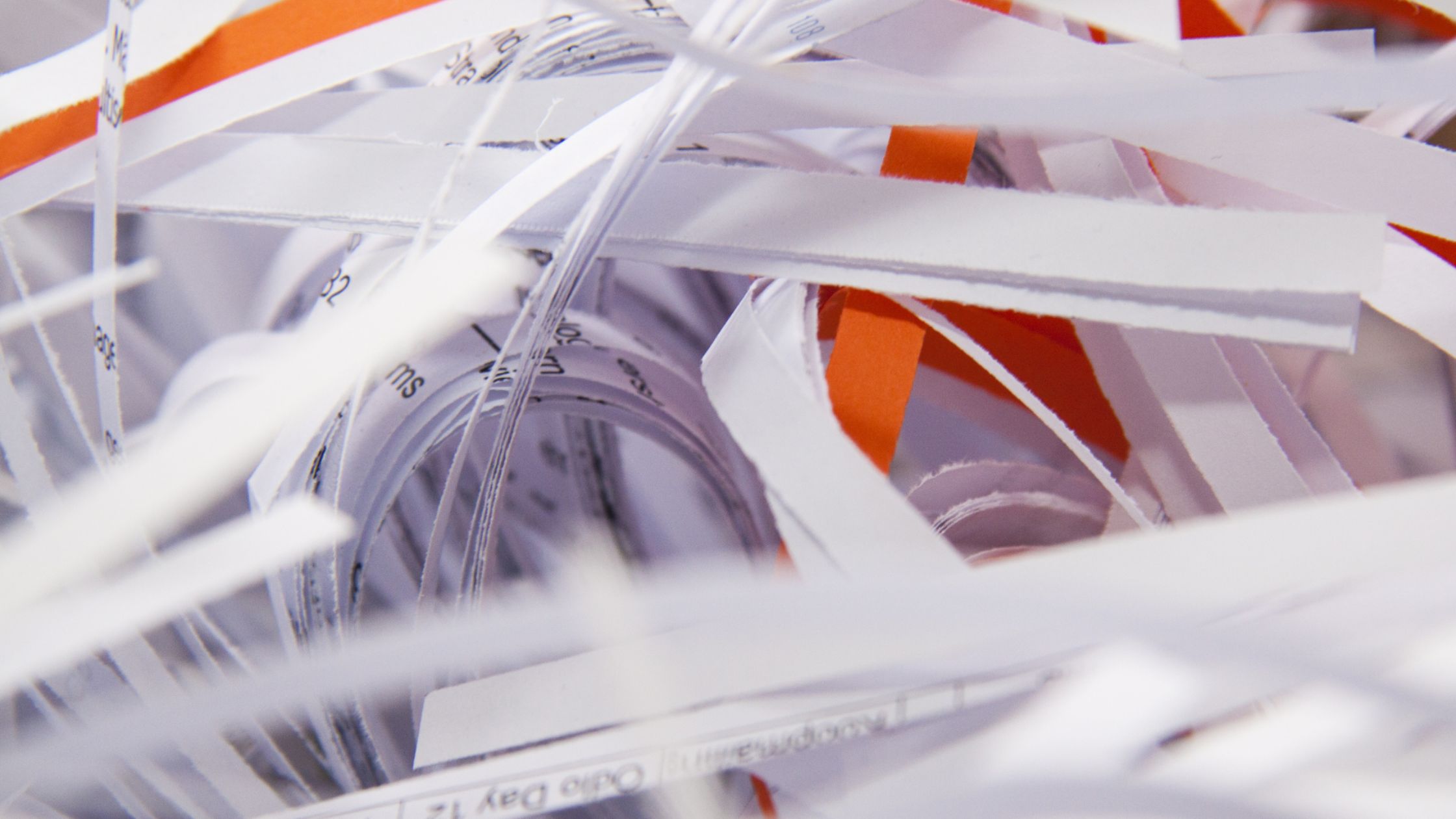 Close-up of shredded paper strips in white and orange, creating a chaotic, tangled texture. The image evokes a sense of clutter and disorder.